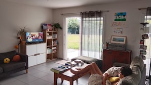 A living room with a sofa, a coffee table, a TV, a bookshelf, and a sliding glass door leading to a garden. - Homestay (Chambéry)