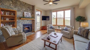 A living room with a stone fireplace, a ceiling fan, and a large window.