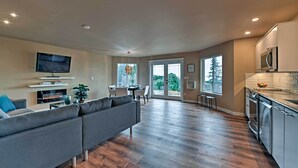A modern living room with a grey sofa, a flat-screen TV, and a fireplace. The kitchen has stainless steel appliances and a large window. - Airy Home Near Anacortes w/ San Juan de Fuca Views (Anacortes)