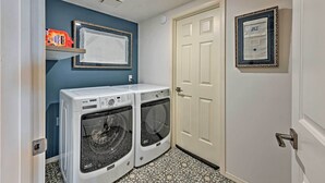 A laundry room with two front-loading washing machines, a shelf with laundry detergent, a framed calendar, and a door. - Airy Home Near Anacortes w/ San Juan de Fuca Views (Anacortes)