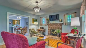 A living room with a fireplace, red upholstered chairs, a dining area with a table and chairs, and a kitchen visible through an open doorway. - Restored Mars Hill Home on 1 Acre: Walk to Town! (Mars Hill)
