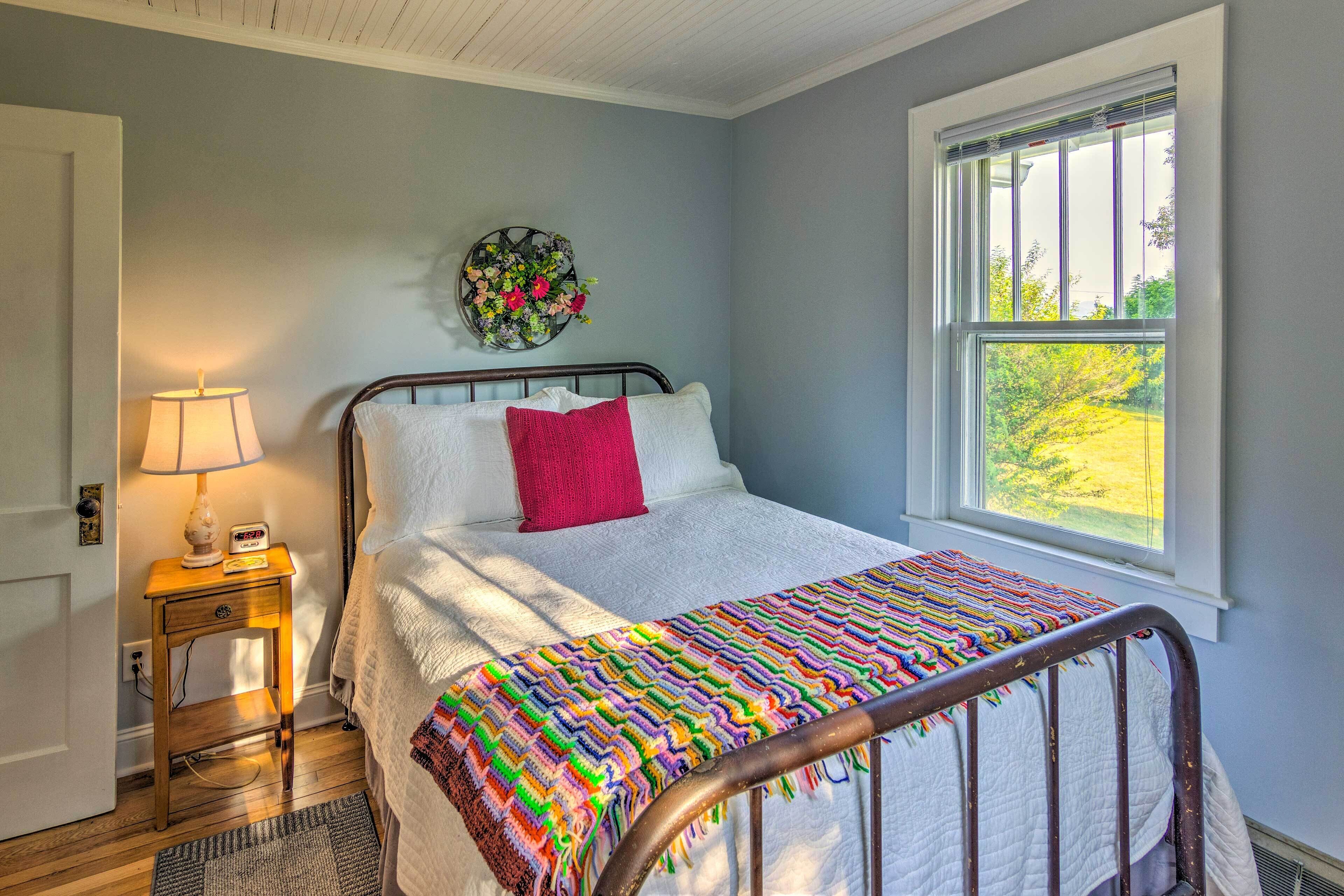 A bedroom with a bed, a nightstand with a lamp, a wall clock, a window with a view of greenery, and a decorative wall hanging.