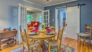 A dining area with a wooden table set for four, surrounded by wooden chairs. There is a chandelier above the table, and a white door with a sliding barn door to the right. - Restored Mars Hill Home on 1 Acre: Walk to Town! (Mars Hill)