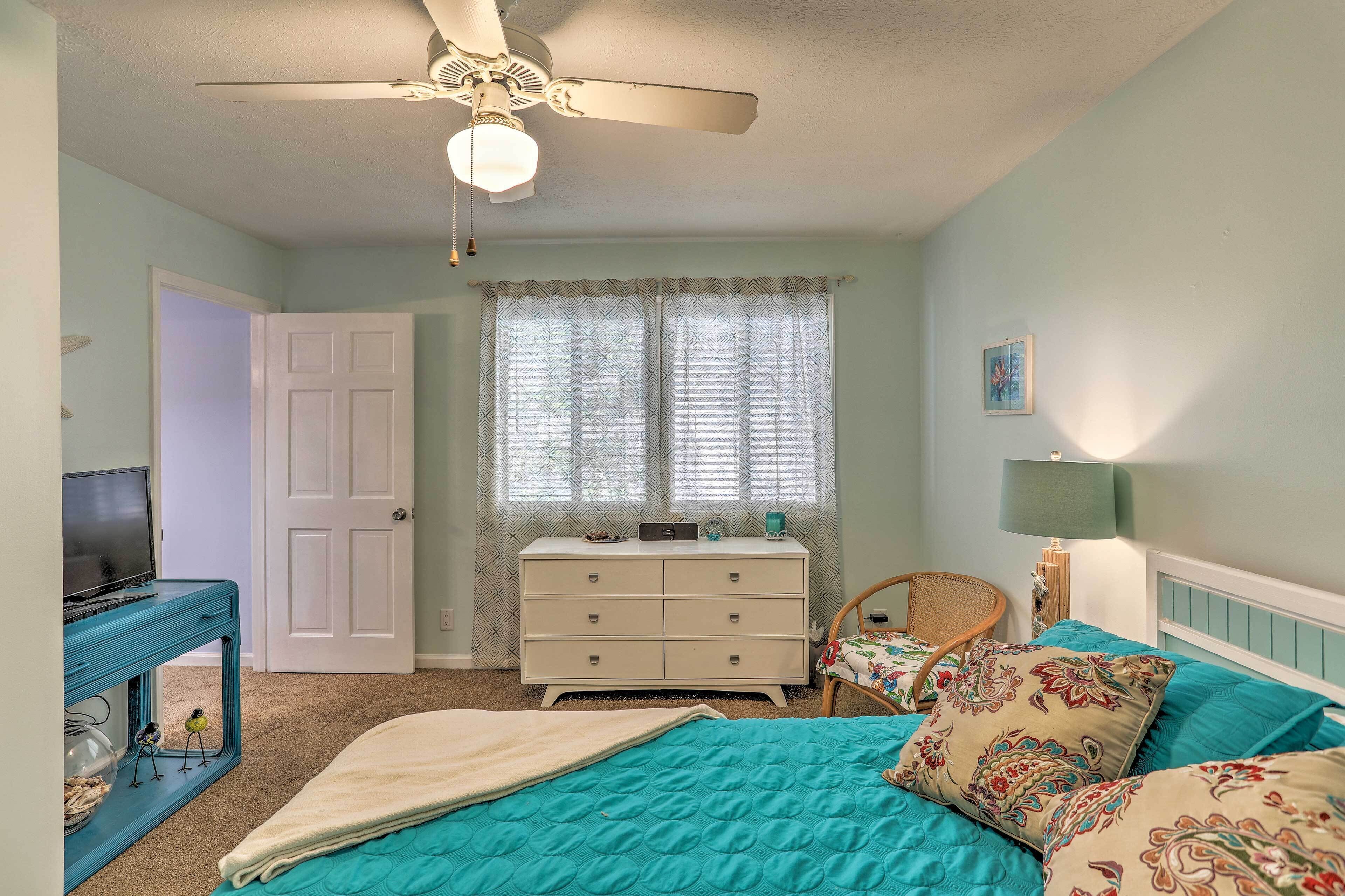 A bedroom with a bed, a dresser, a chair, a desk with a TV, a ceiling fan, and a window with curtains.