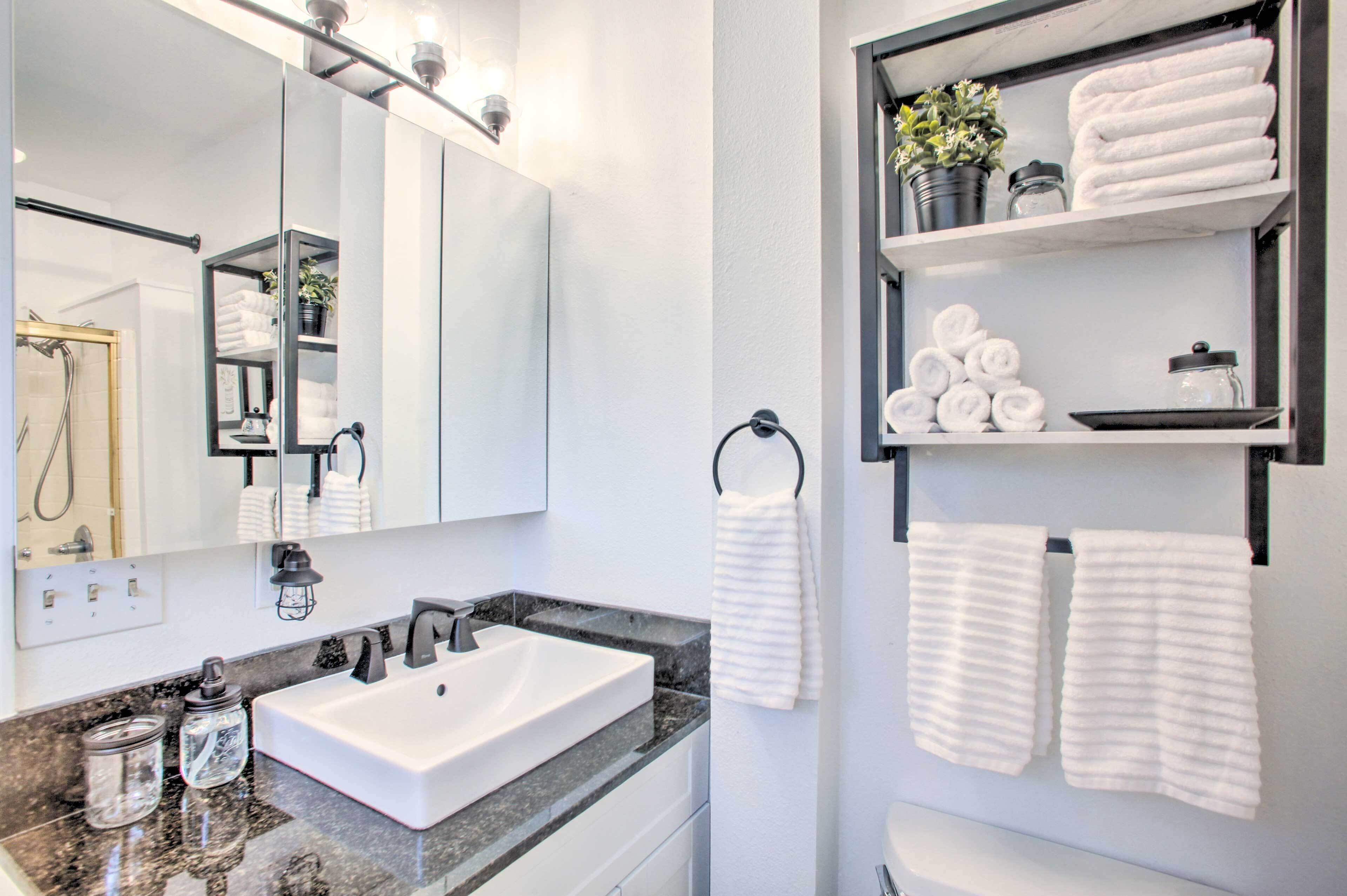 A modern bathroom with a large mirror, white sink, and marble countertop. There are shelves with towels and toiletries.