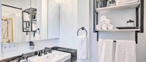 A modern bathroom with a large mirror, white sink, and marble countertop. There are shelves with towels and toiletries.
