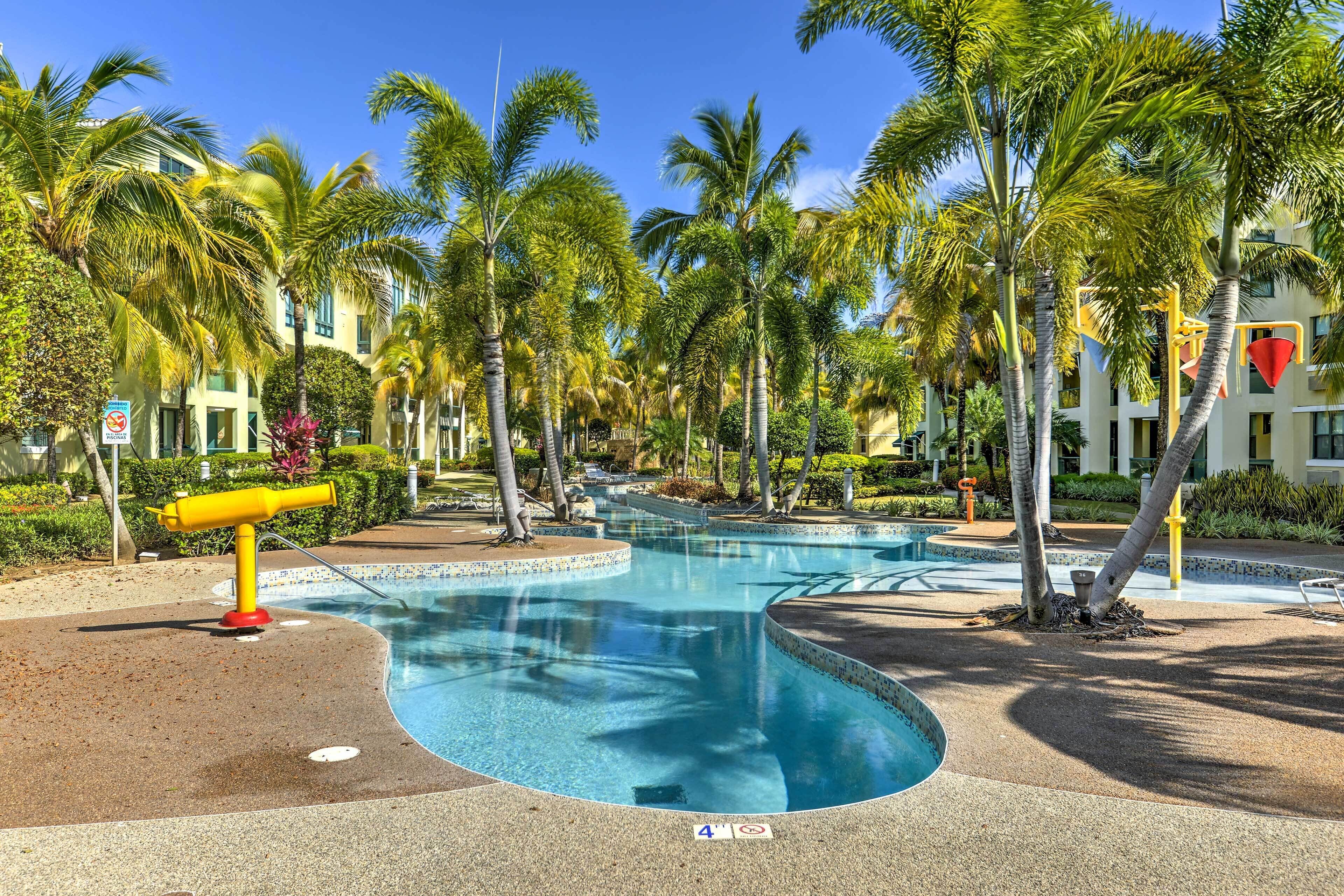 A swimming pool with a curved design, surrounded by palm trees and a building with a yellow facade.