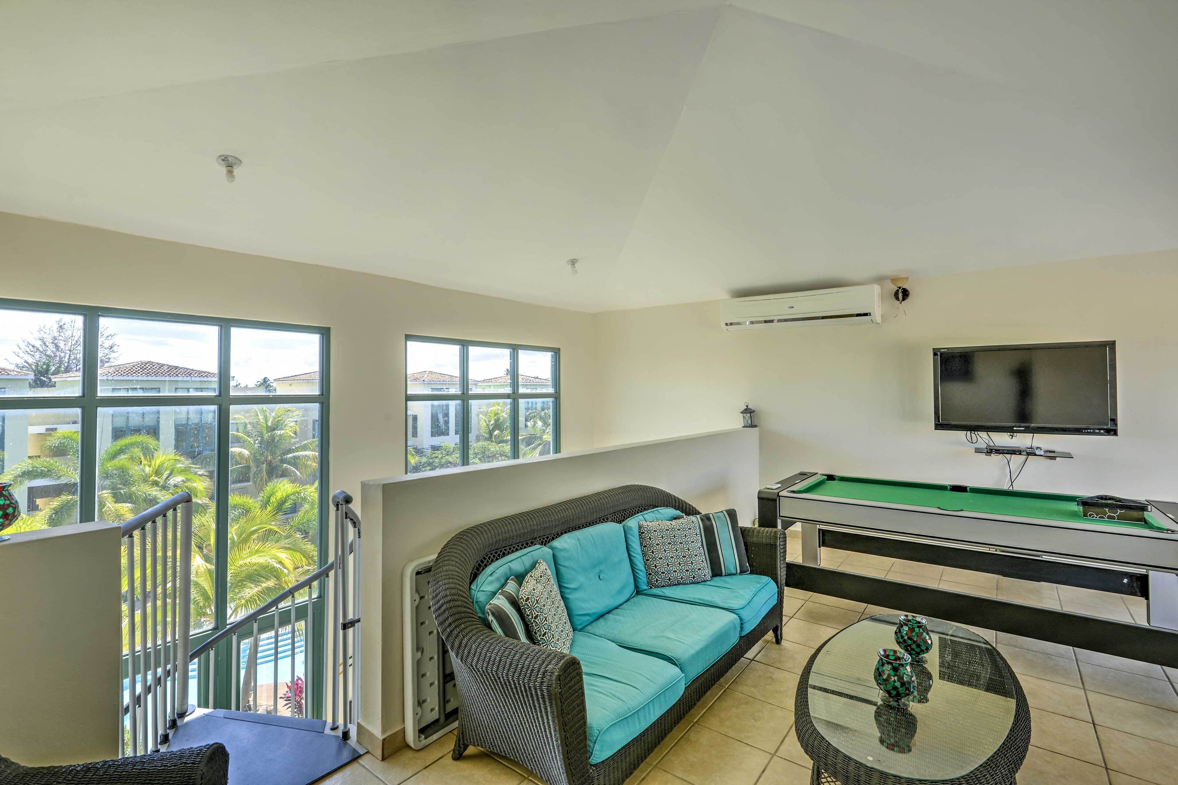 Una habitación con sofá, mesa de billar, televisor y balcón con vista a palmeras.
