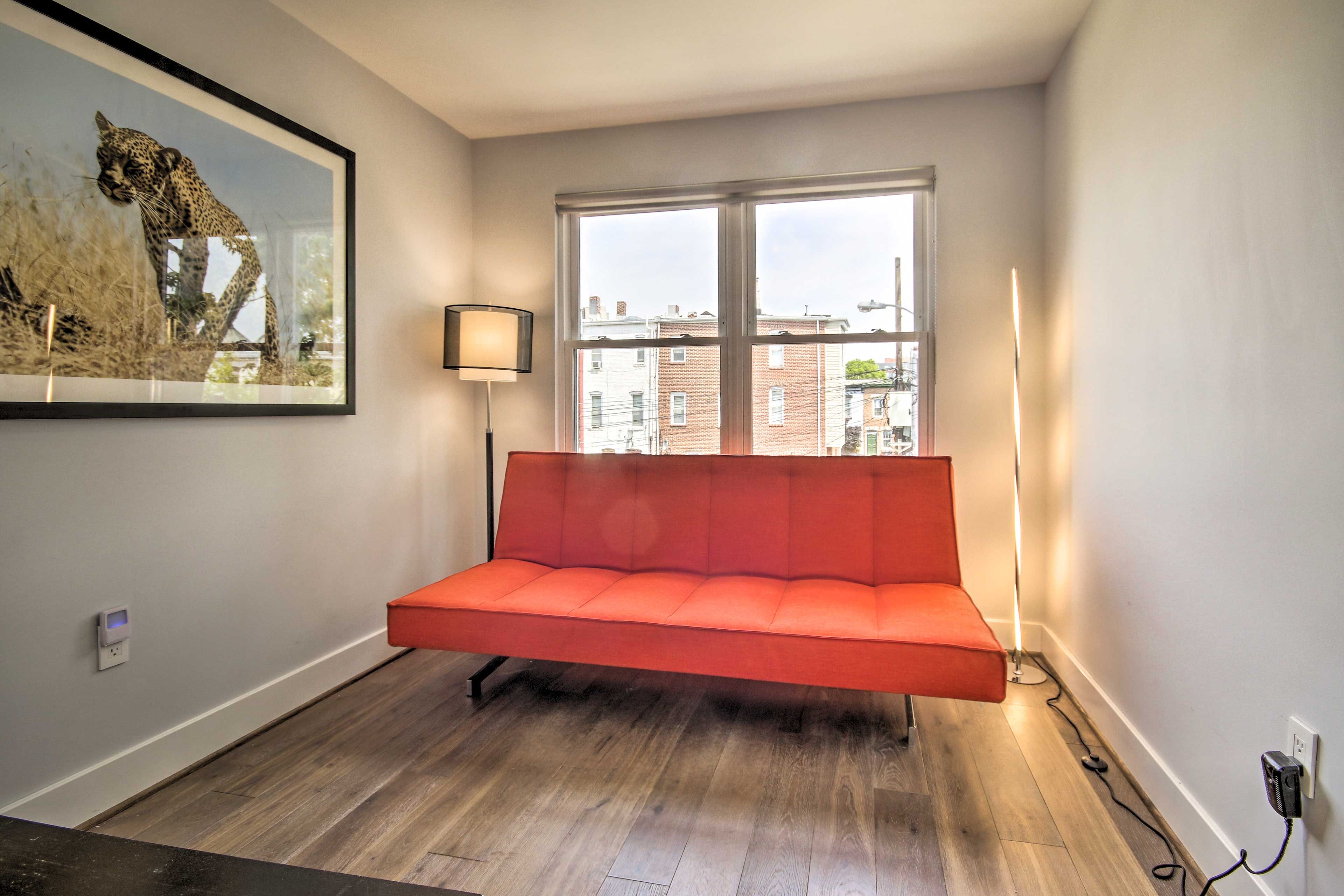 Een moderne kamer met een rode bank, een ingelijst fototoestel van dieren in het wild aan de muur, en een groot raam met uitzicht op gebouwen.