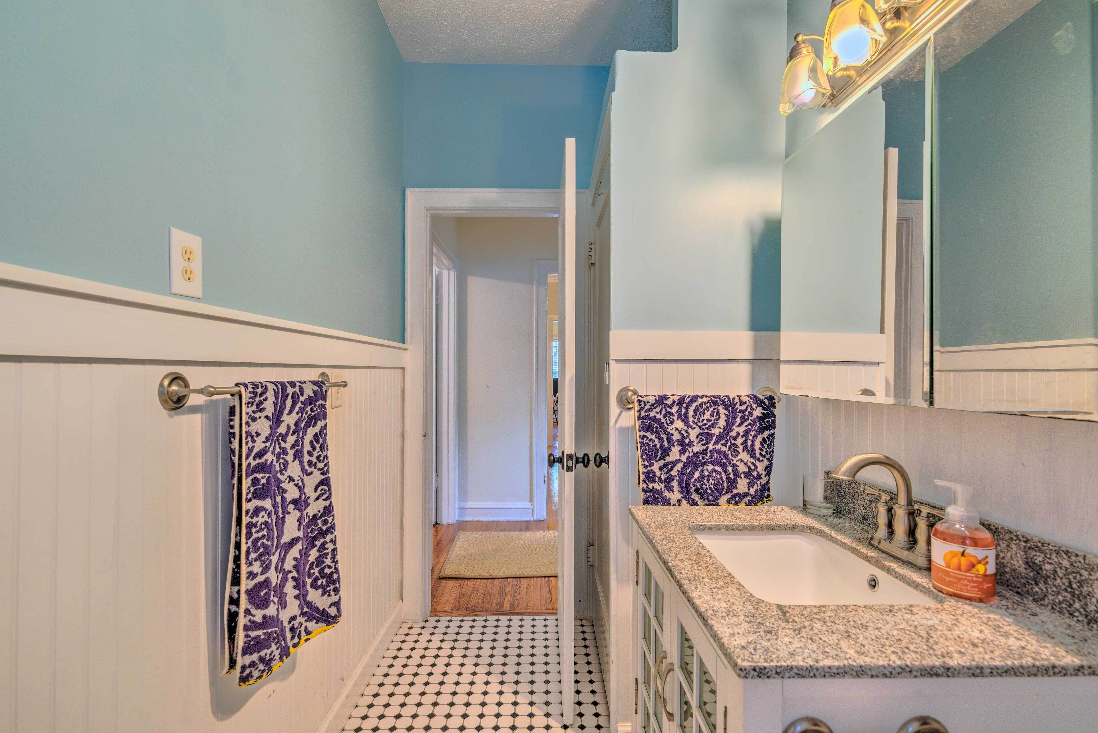 A narrow bathroom with a white vanity, a sink, and a mirror. A towel hangs on the wall, and a door leads to another room.
