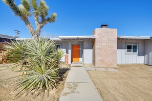 A single-story house with a brick chimney, a concrete pathway leading to an orange door, and a large yucca plant in the foreground. - Joshua Tree Vacation Home w/ Private Hot Tub! (Joshua Tree)
