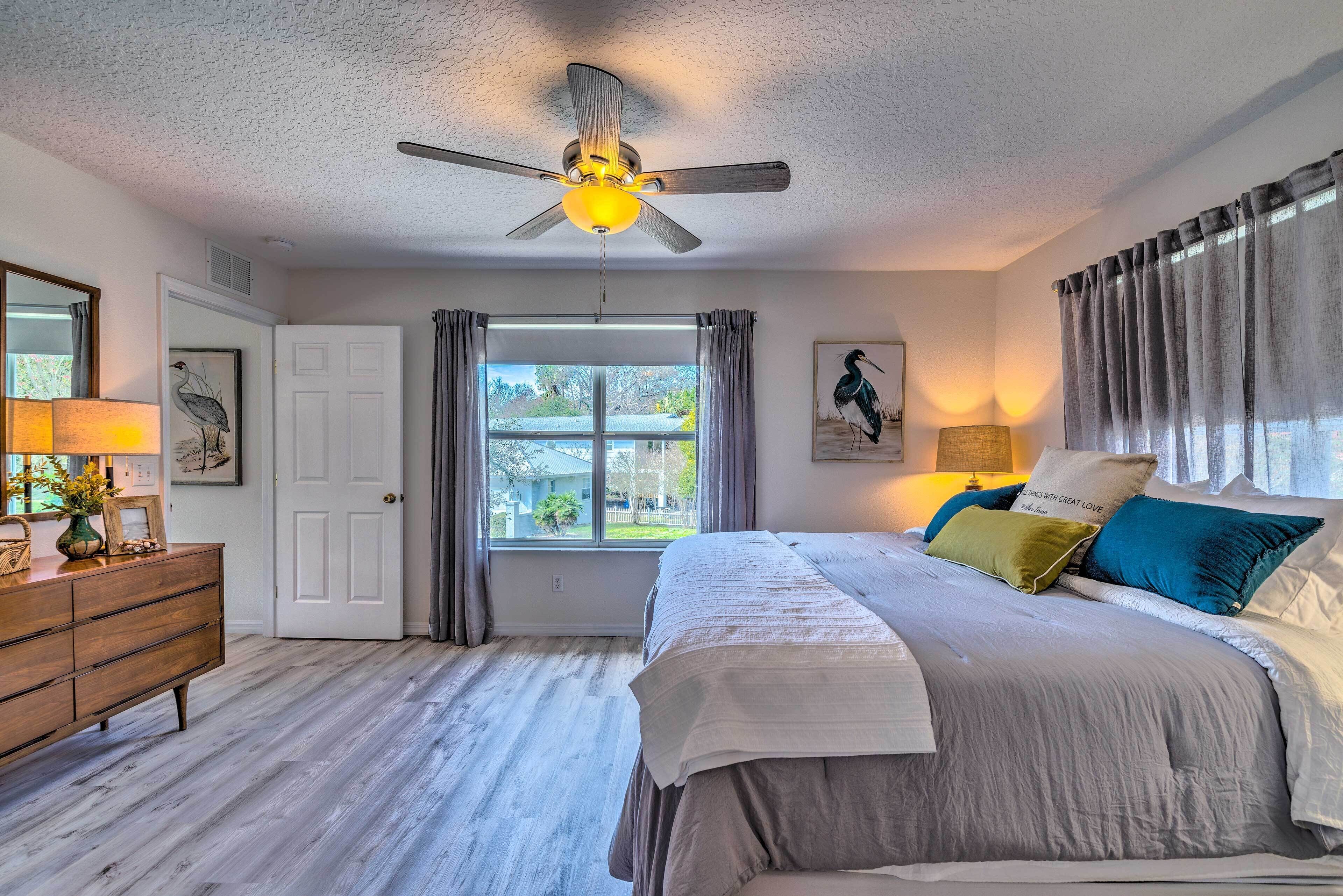 A bedroom with a bed, a ceiling fan, a dresser, a window with curtains, and a view of the outside.