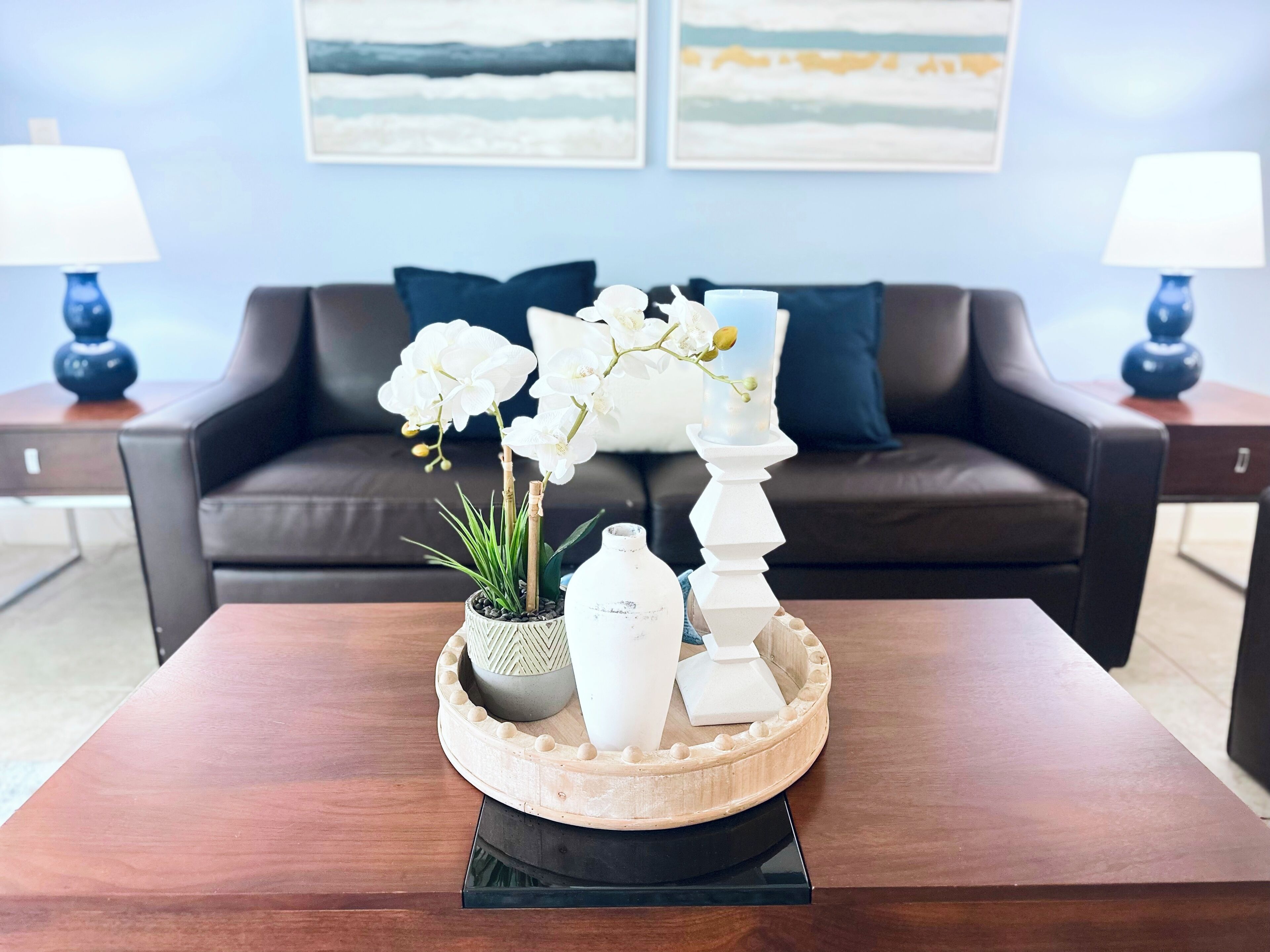 A modern living room with a dark brown leather sofa, two blue lamps, a wooden coffee table with a decorative tray, white orchids, and a tall white candle holder.