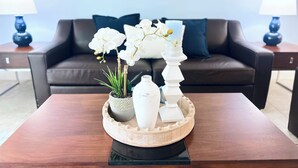 Een moderne woonkamer met een donkerbruine leren bank, twee blauwe lampen, een houten salontafel met een decoratief dienblad, witte orchideeën en een hoge witte kandelaar.
