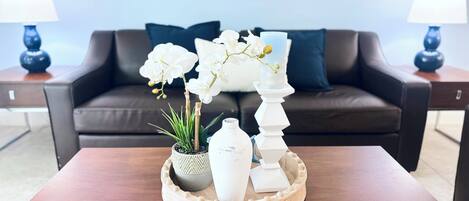A modern living room with a dark brown leather sofa, two blue lamps, a wooden coffee table with a decorative tray, white orchids, and a tall white candle holder.