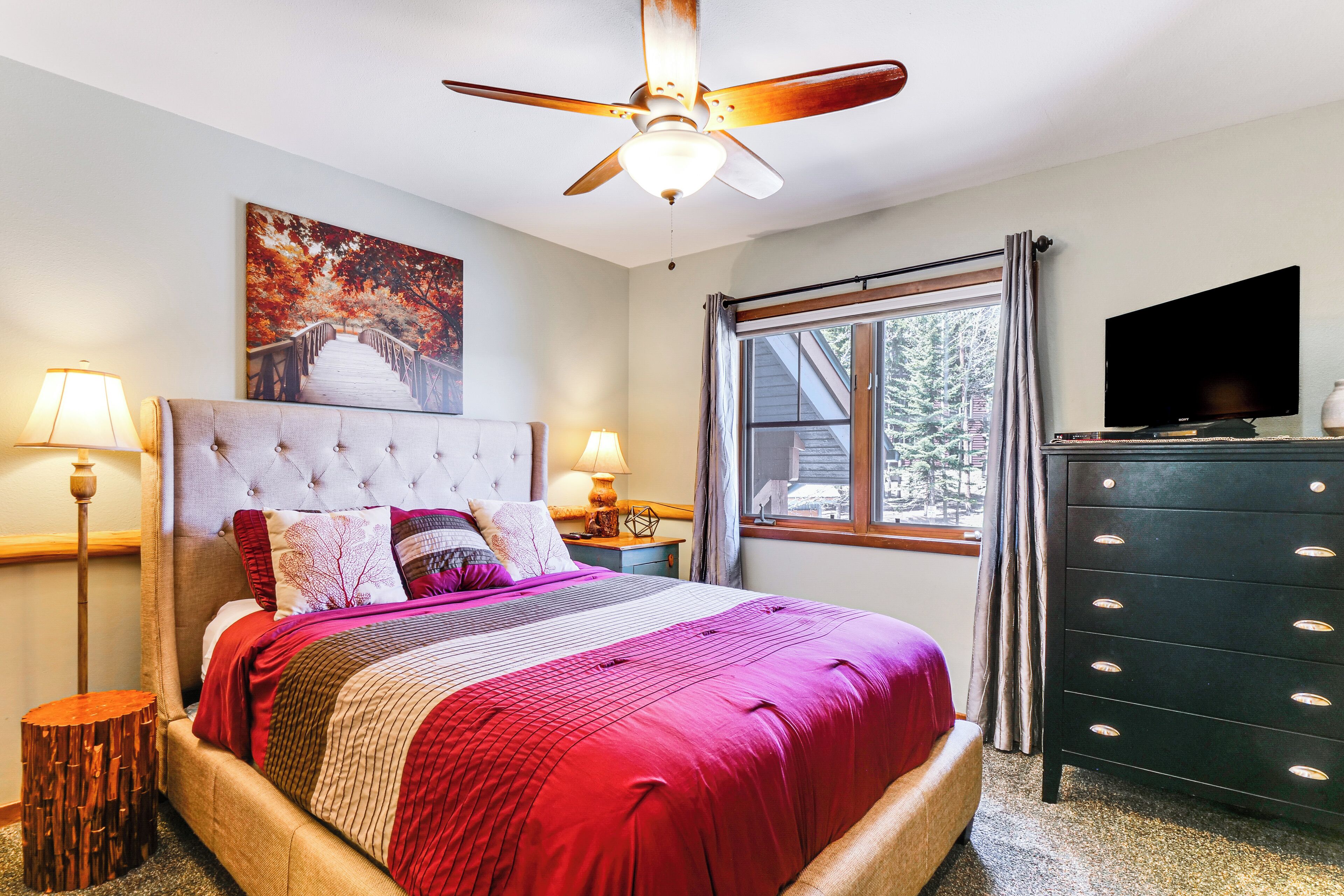 Une chambre avec un grand lit, un ventilateur de plafond, une télévision et une fenêtre avec des rideaux.