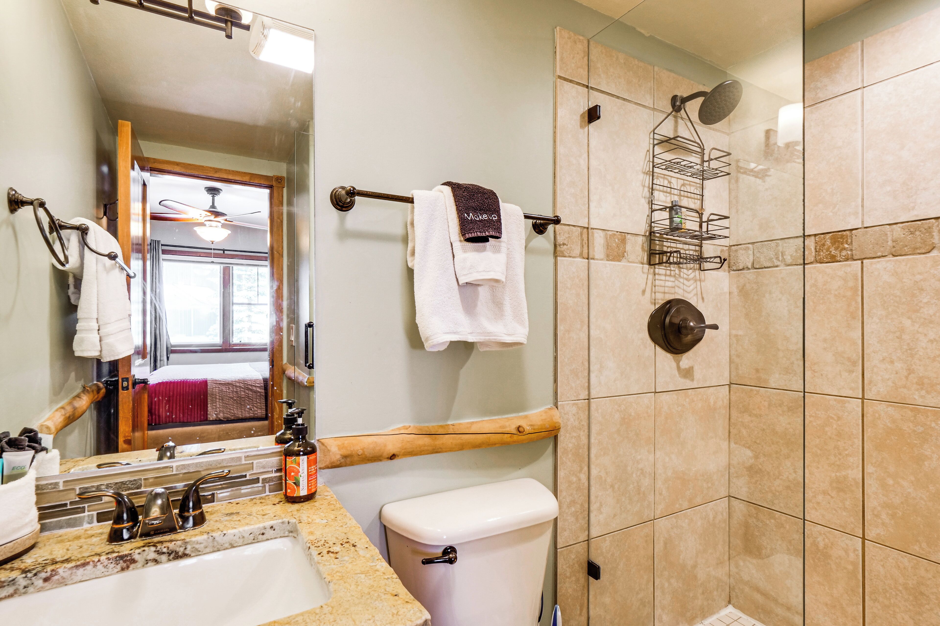 Une salle de bain avec un lavabo, des toilettes et une douche. On y trouve un miroir, un porte-serviettes et une étagère en bois.