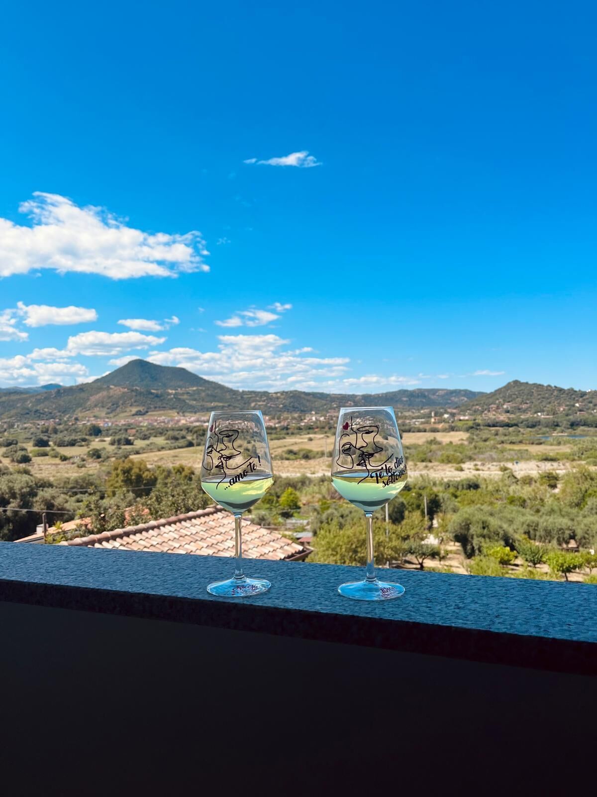 Dos copas de vino con diseños de rostros en un balcón con vistas a un paisaje pintoresco con montañas y cielo despejado.