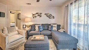 Un salon avec un canapé d’angle gris, un fauteuil beige, un ventilateur de plafond et des œuvres d’art décoratives accrochées au mur.