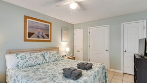 A bedroom with a bed, a ceiling fan, a painting, a nightstand, a lamp, and a TV. - 1st-floor Resort Condo in PCB - Walk to Beach! (Panama City Beach)