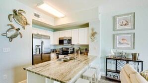 A modern kitchen with white cabinets, a marble countertop, and a stainless steel refrigerator. There are two wall-mounted turtle decorations and framed artwork above the counter. - Beachfront PCB Condo w/ Ocean View & Pool Access! (Panama City Beach)