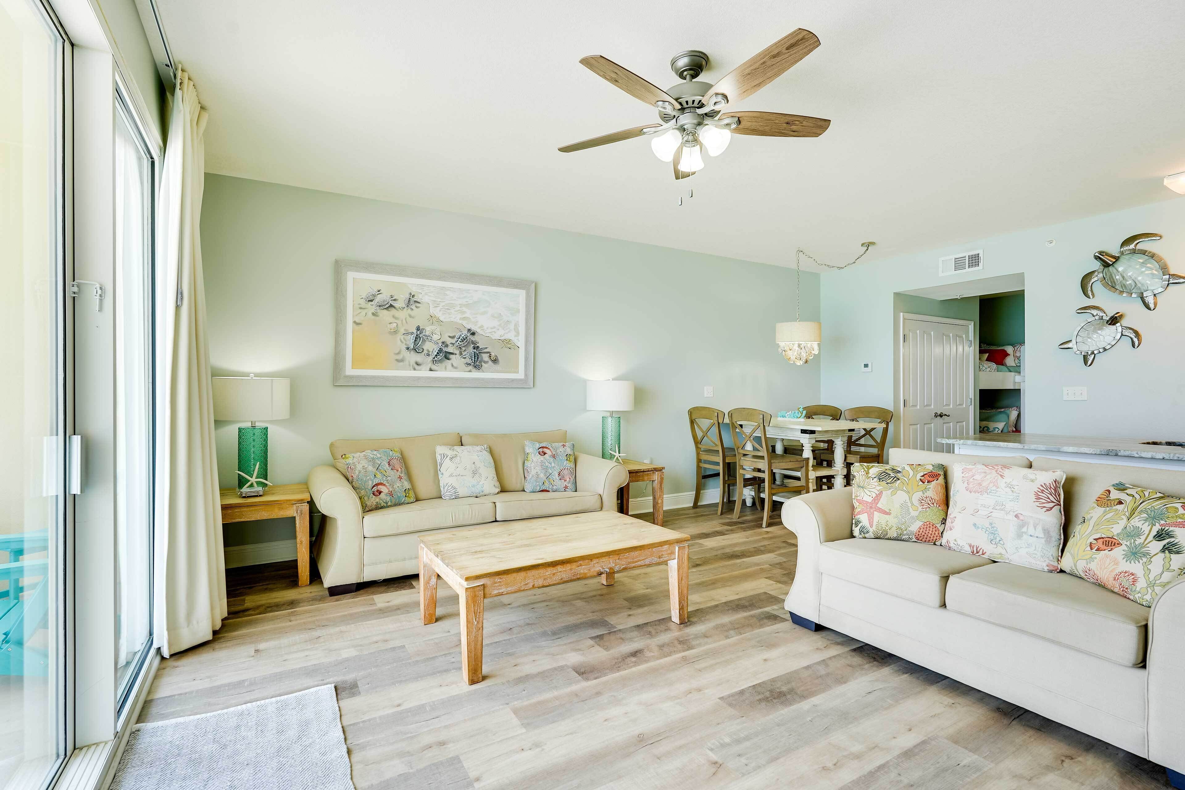 A living room with a wooden coffee table, beige sofas, a ceiling fan, and a dining area with chairs and a table.