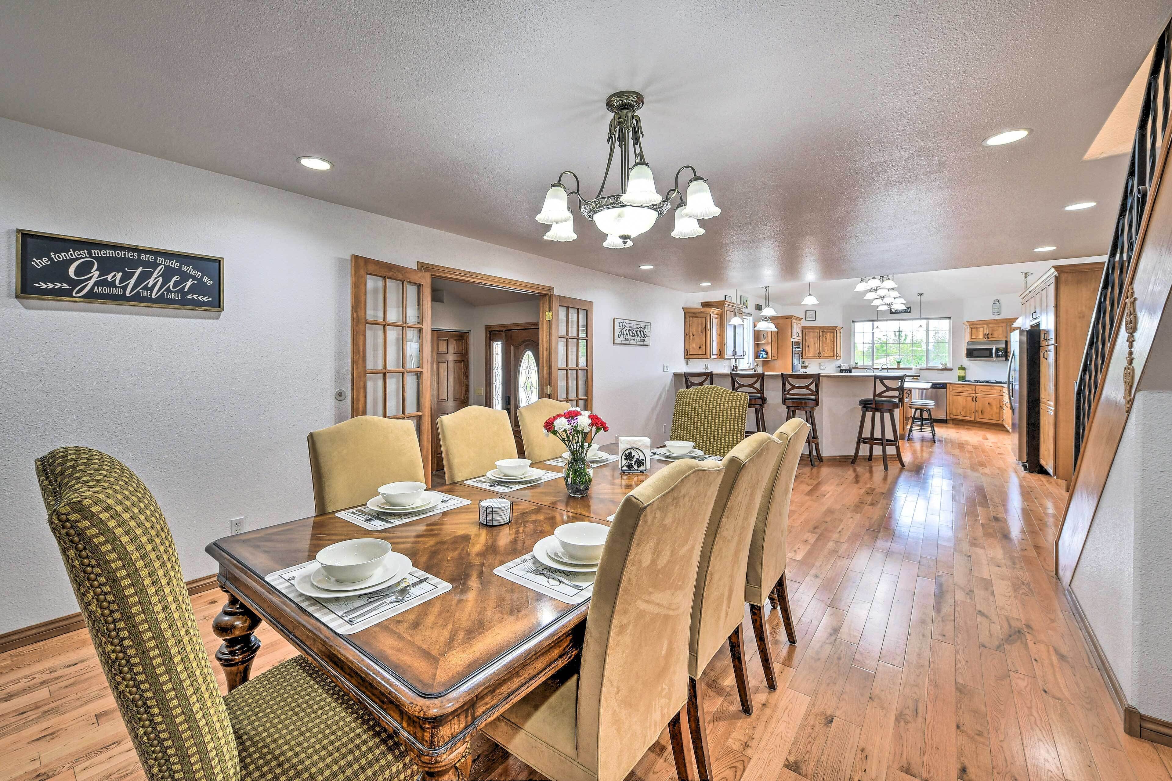 A dining area with a wooden table set for four, surrounded by upholstered chairs. A chandelier hangs above the table, and a kitchen is visible in the background.