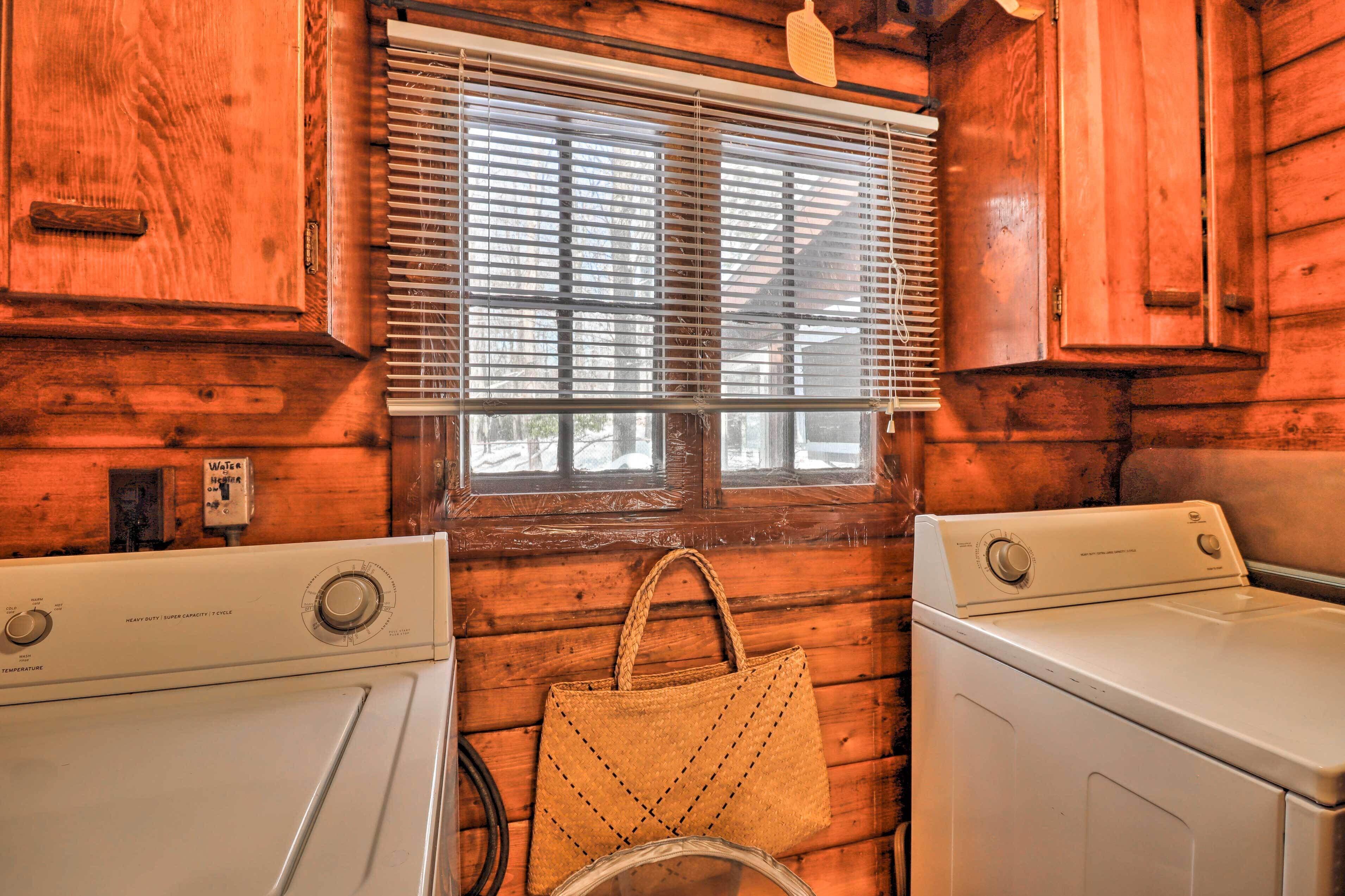 A laundry room with two washing machines, a window with blinds, and a woven bag hanging on the wall.