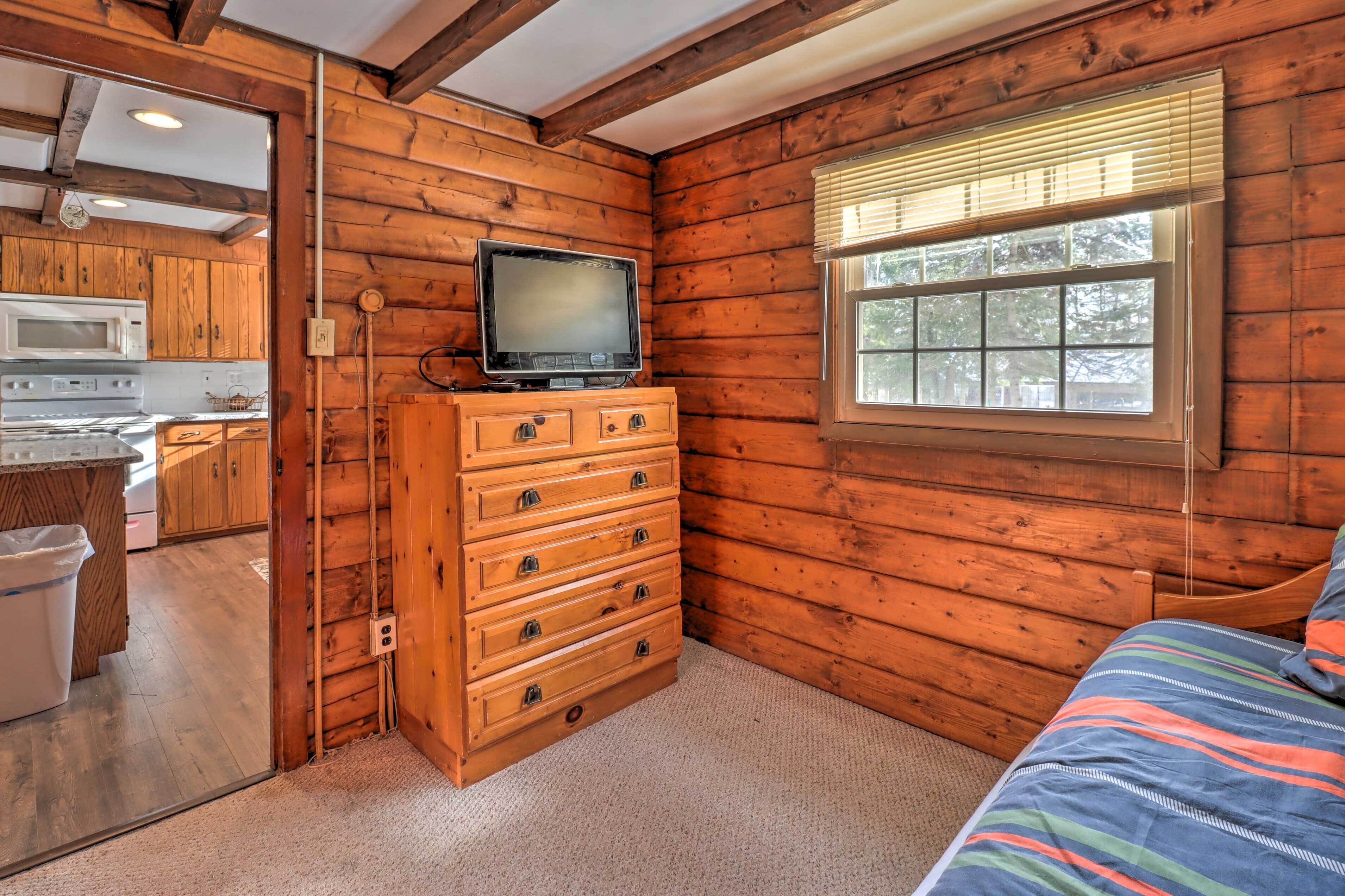 Una habitación tipo cabaña de troncos con tocador de madera, televisor, ventana con persianas y cama con una manta colorida.