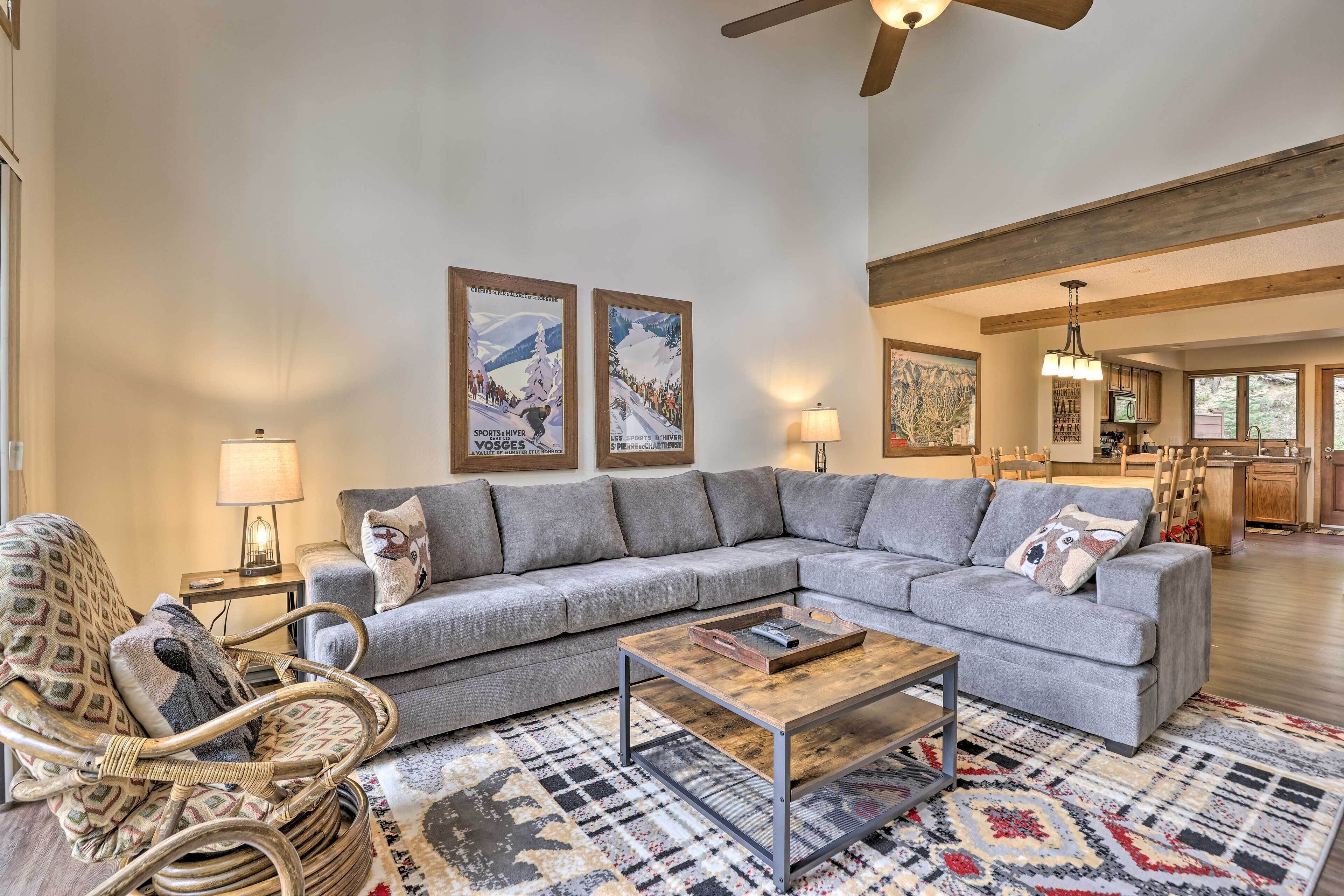 A spacious living room with a grey sectional sofa, a wooden coffee table, and a patterned rug. There is a wicker chair with a cushion, a lamp, and framed pictures on the wall. A kitchen area is visible in the background.