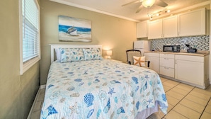 A bedroom with a bed, a nightstand, a chair, a ceiling fan, and a kitchen area with cabinets and a microwave. - Beachfront PCB Studio: Pool, Patio & Deck Access! (Panama City Beach)