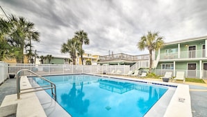 A swimming pool with a white fence, palm trees, and a building with balconies in the background. - Beachfront PCB Studio: Pool, Patio & Deck Access! (Panama City Beach)