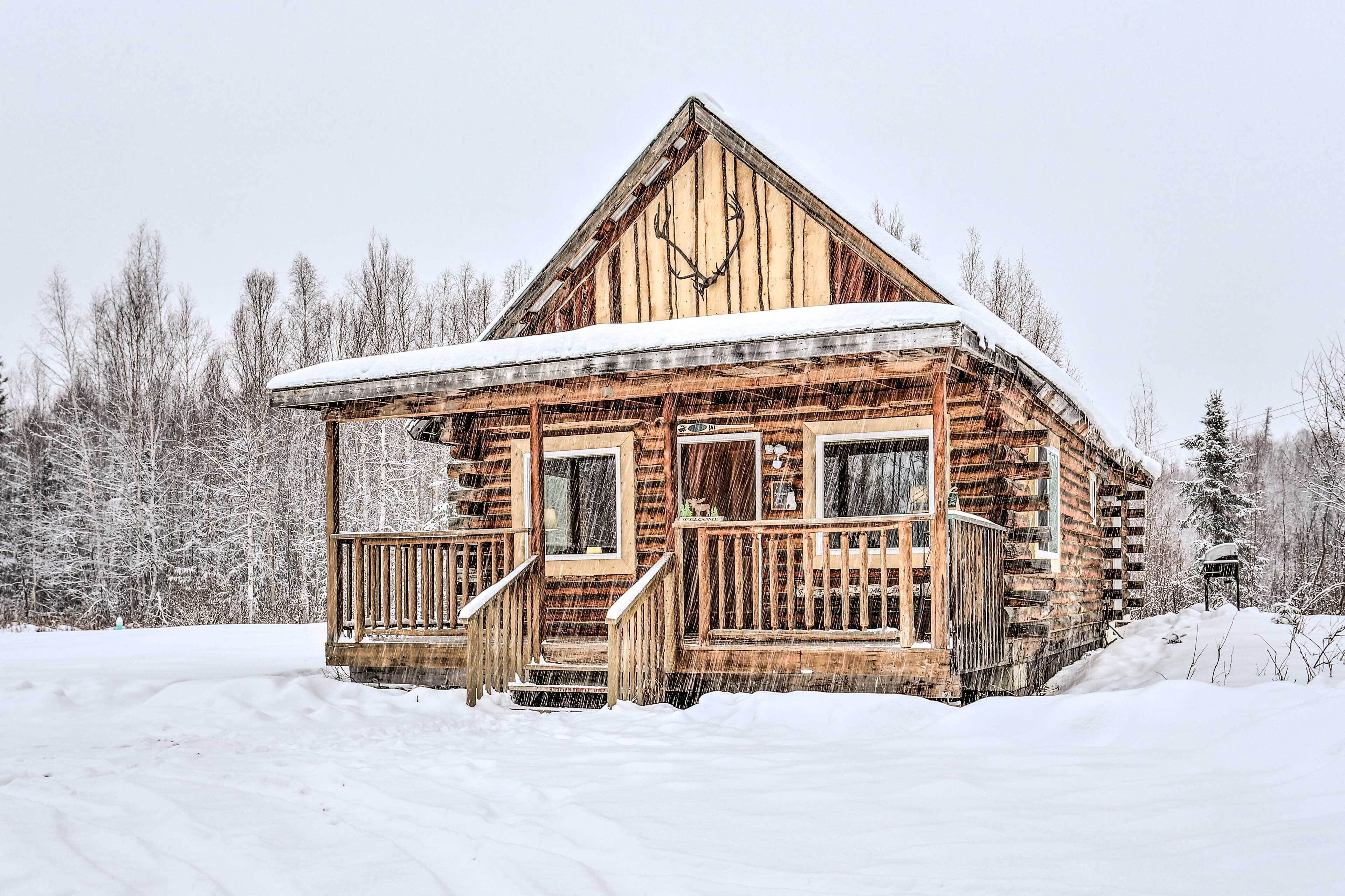 Una cabaña rústica de troncos con un porche de madera y un techo cubierto de nieve, rodeada por un paisaje nevado.