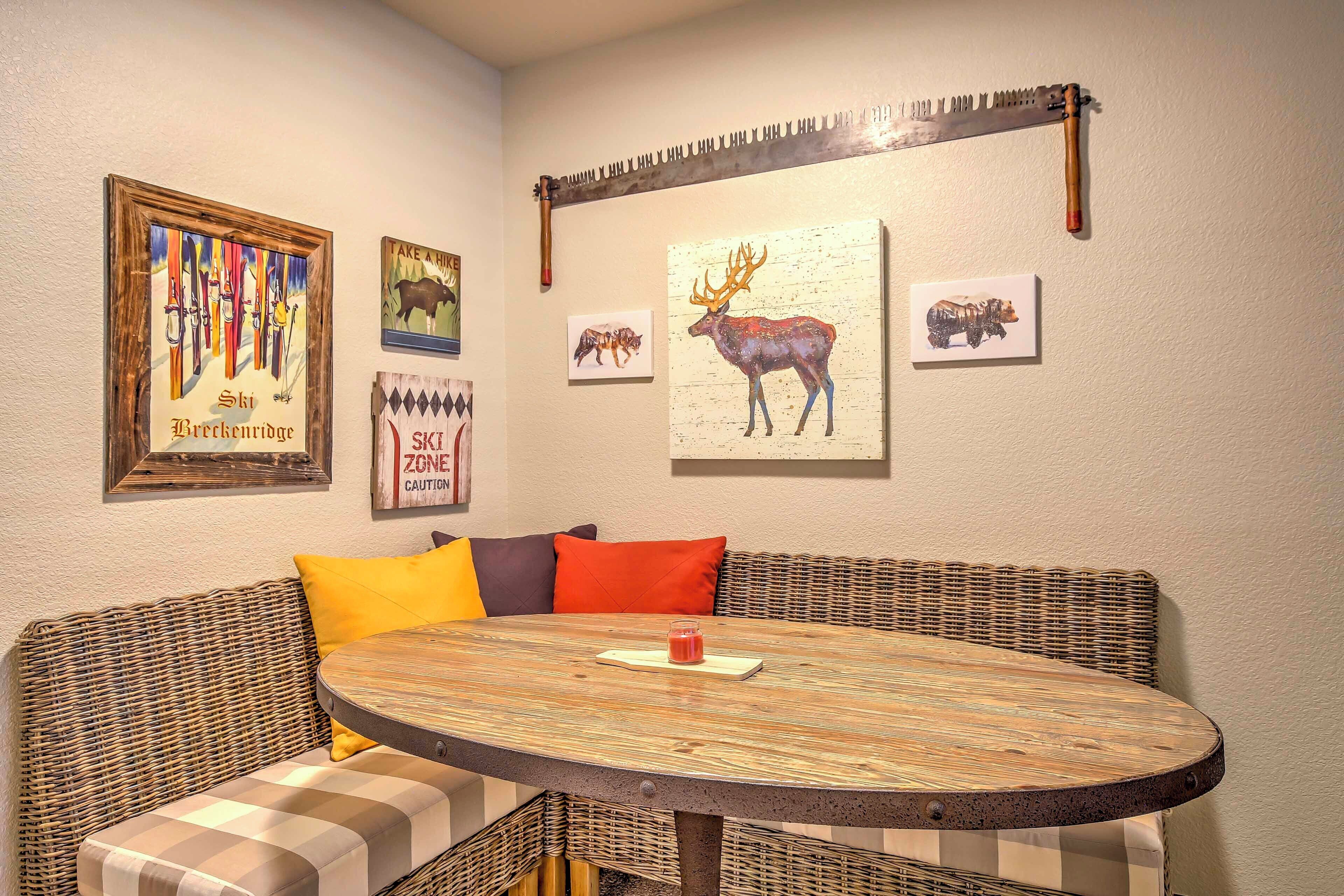 Een gezellige zithoek met een ronde tafel, rieten meubels en wanddecoratie, waaronder een schilderij van een hert en kunstwerken met een wintersportthema.