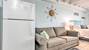 A room with a white refrigerator, a beige sofa with a cushion, a wall-mounted sun decoration, and a bed with a blue headboard. - Panama City Beach Studio w/ Pool & Beach Access (Panama City Beach)