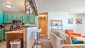 Une cuisine moderne avec des armoires de couleur turquoise, un four à micro-ondes en acier inoxydable et un tabouret de bar. Le salon est doté d’un canapé beige et d’un fauteuil orange, ainsi que d’une étagère en bois et de tableaux encadrés au mur.