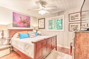 A bedroom with a wooden bed, a ceiling fan, a window with a view of greenery, and framed pictures on the wall. - Redwoods Cabin w/ Hot Tub: Walk to Russian River! (Guerneville)