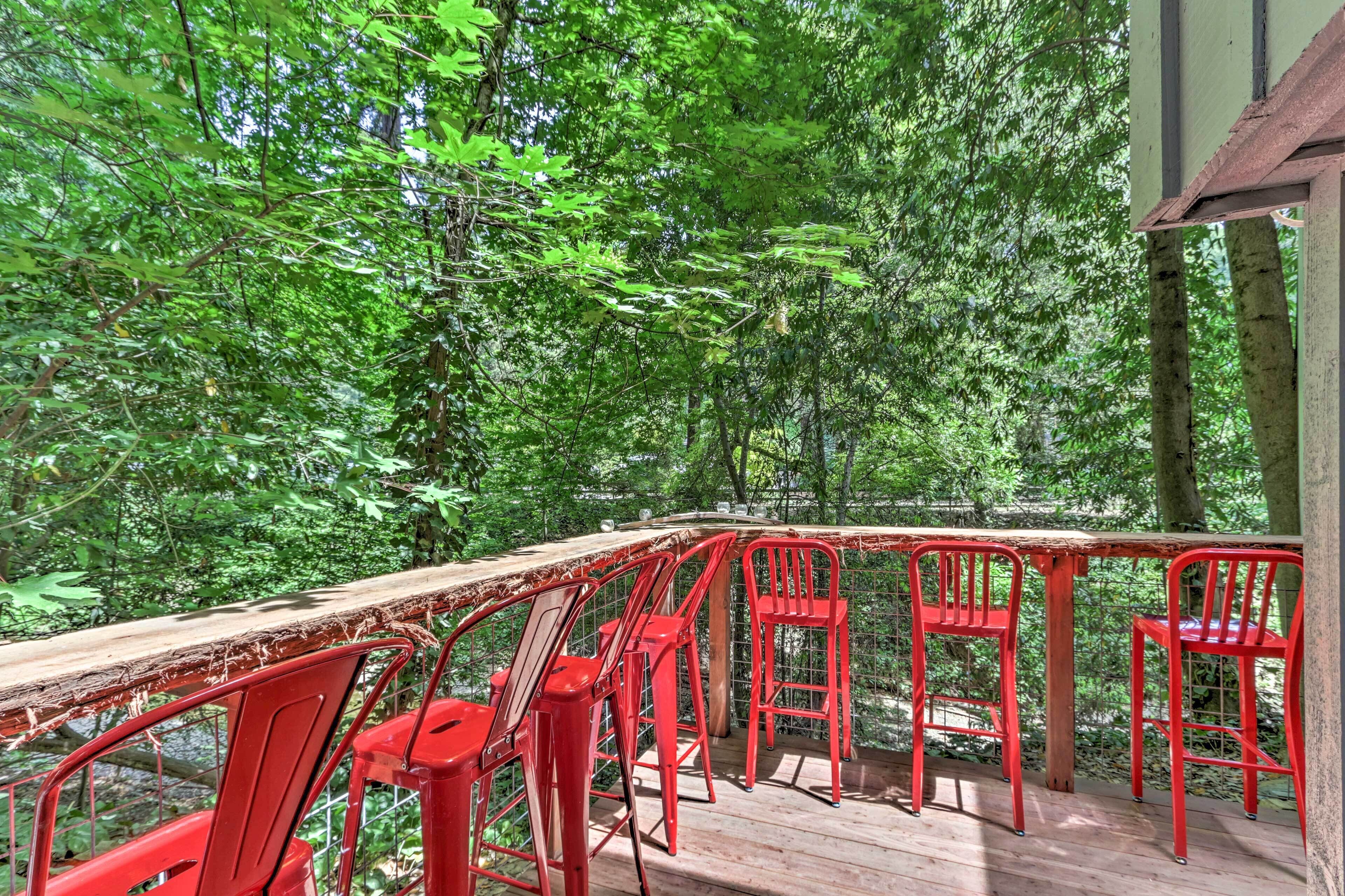 A wooden deck with red metal chairs and a table, surrounded by dense green foliage.