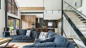 A modern living room with a large sofa set, a glass staircase, and a kitchen in the background. - The Northern Escape HOT TUB & SAUNA! (blue mountain)