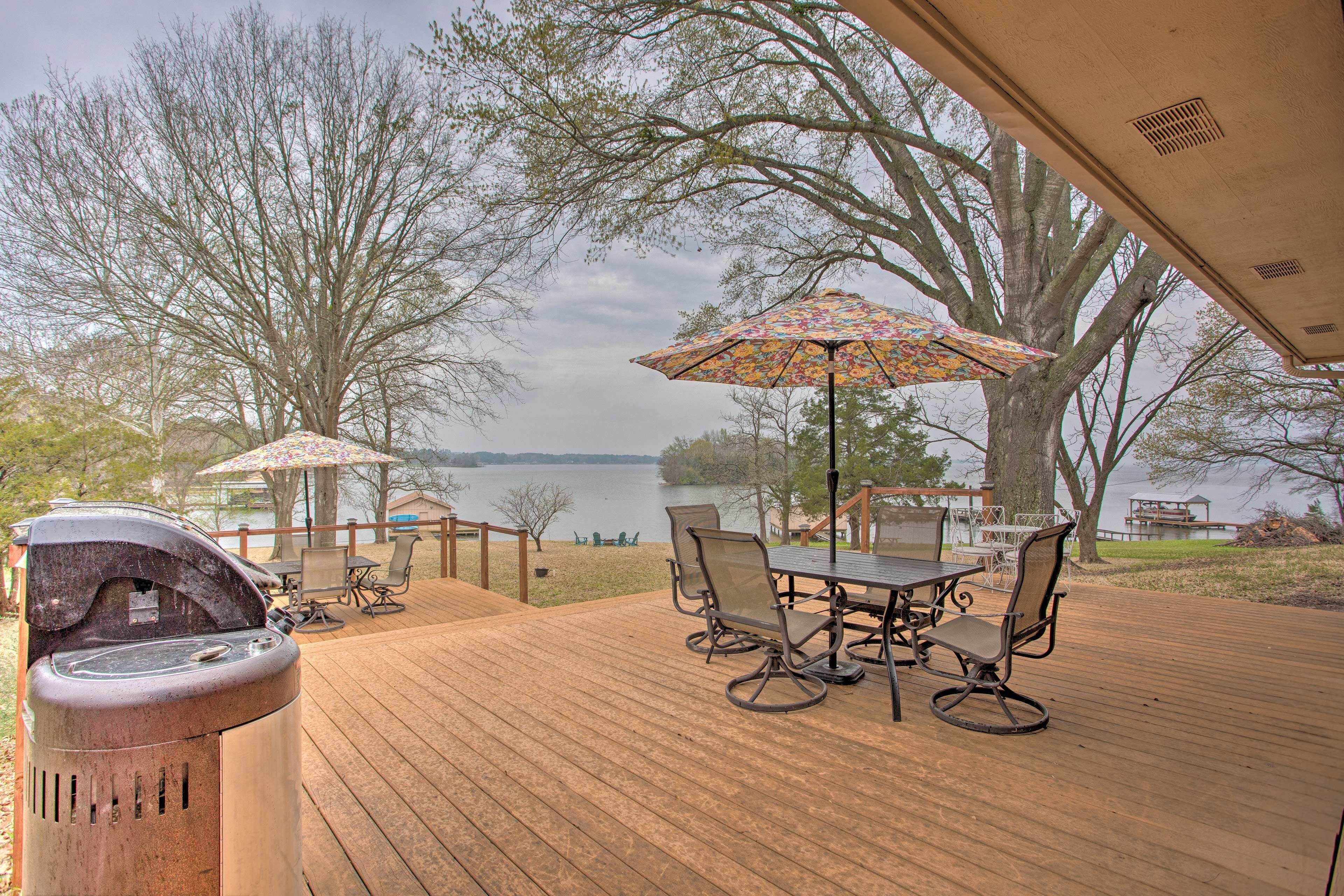 A wooden deck with outdoor furniture, a grill, and umbrellas overlooking a lake.