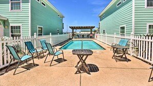 A pool area with lounge chairs and a table, surrounded by a white fence and two buildings with green siding. - Near Port Aransas Beach: Condo w/ Pool Access (Port Aransas)