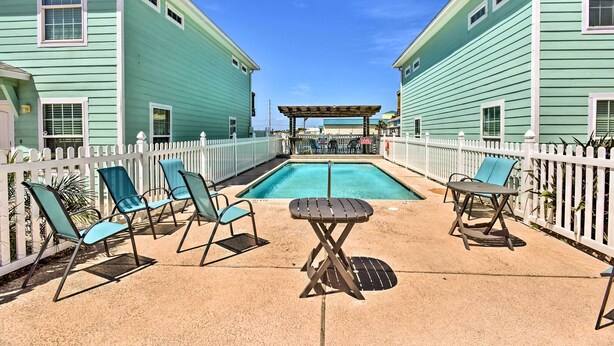 A pool area with lounge chairs and a table, surrounded by a white fence and two buildings with green siding.