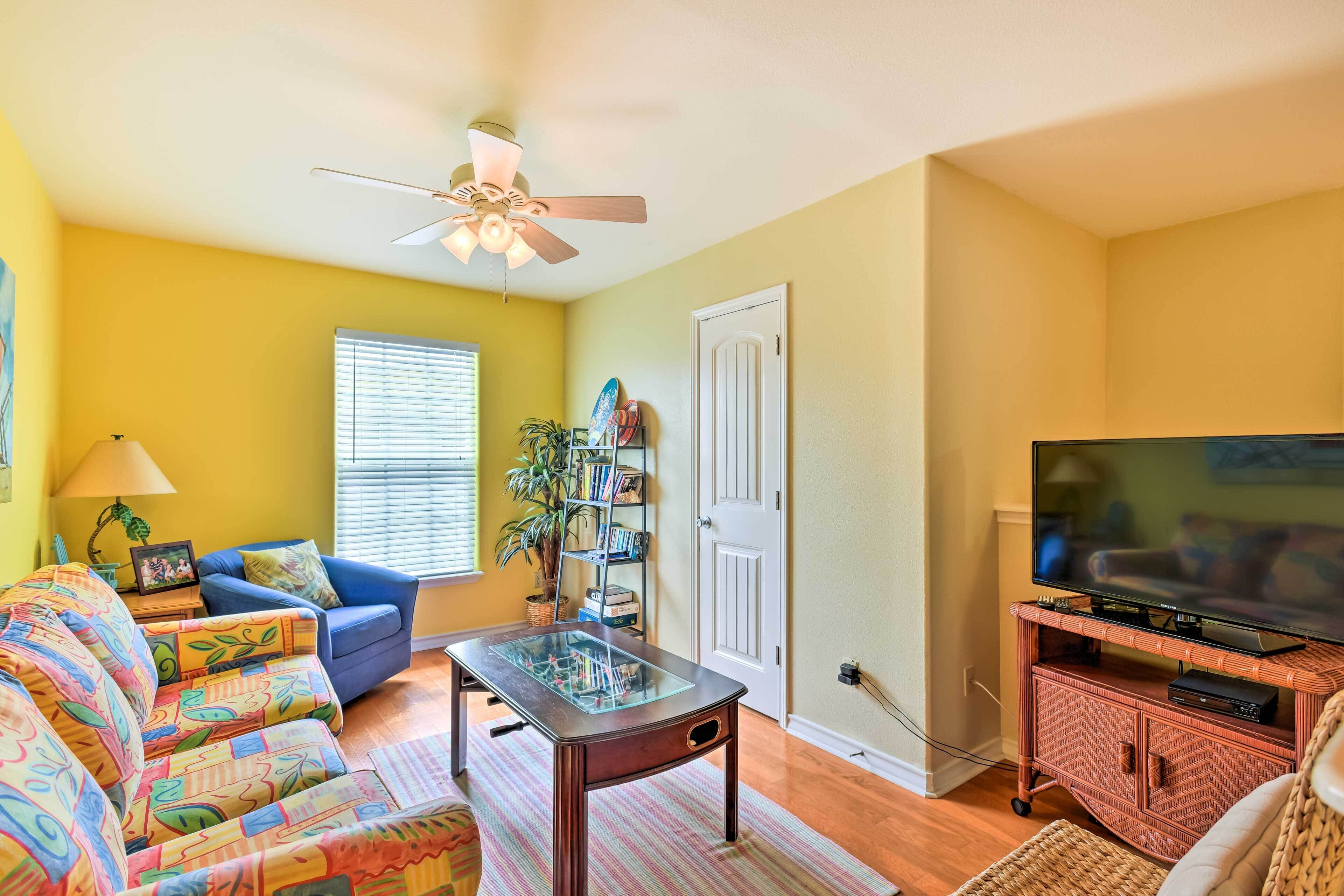A living room with a sofa, a glass-top coffee table, a television on a stand, and a ceiling fan.