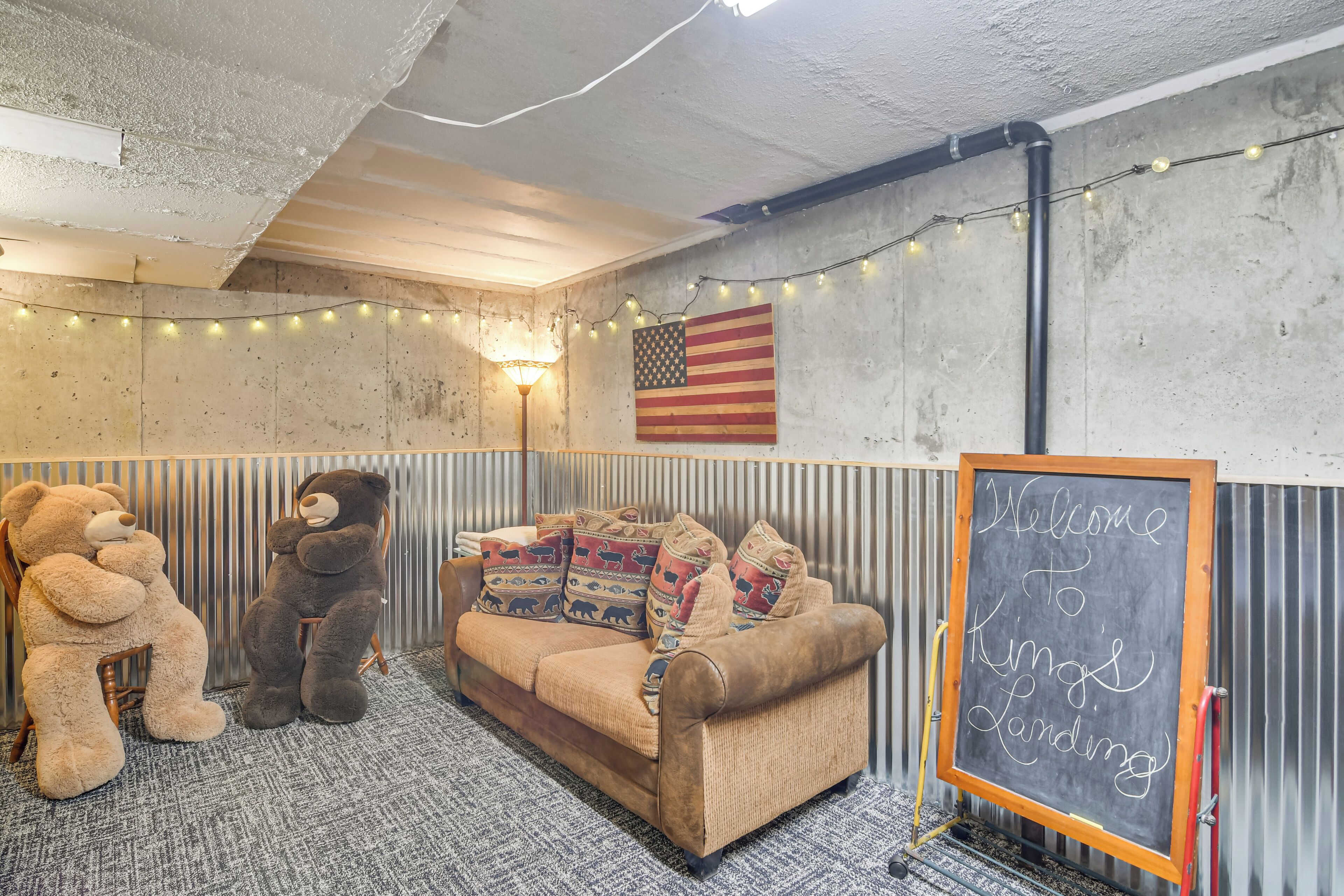 A room with a couch, two large teddy bears, a wall-mounted American flag, and a chalkboard sign.