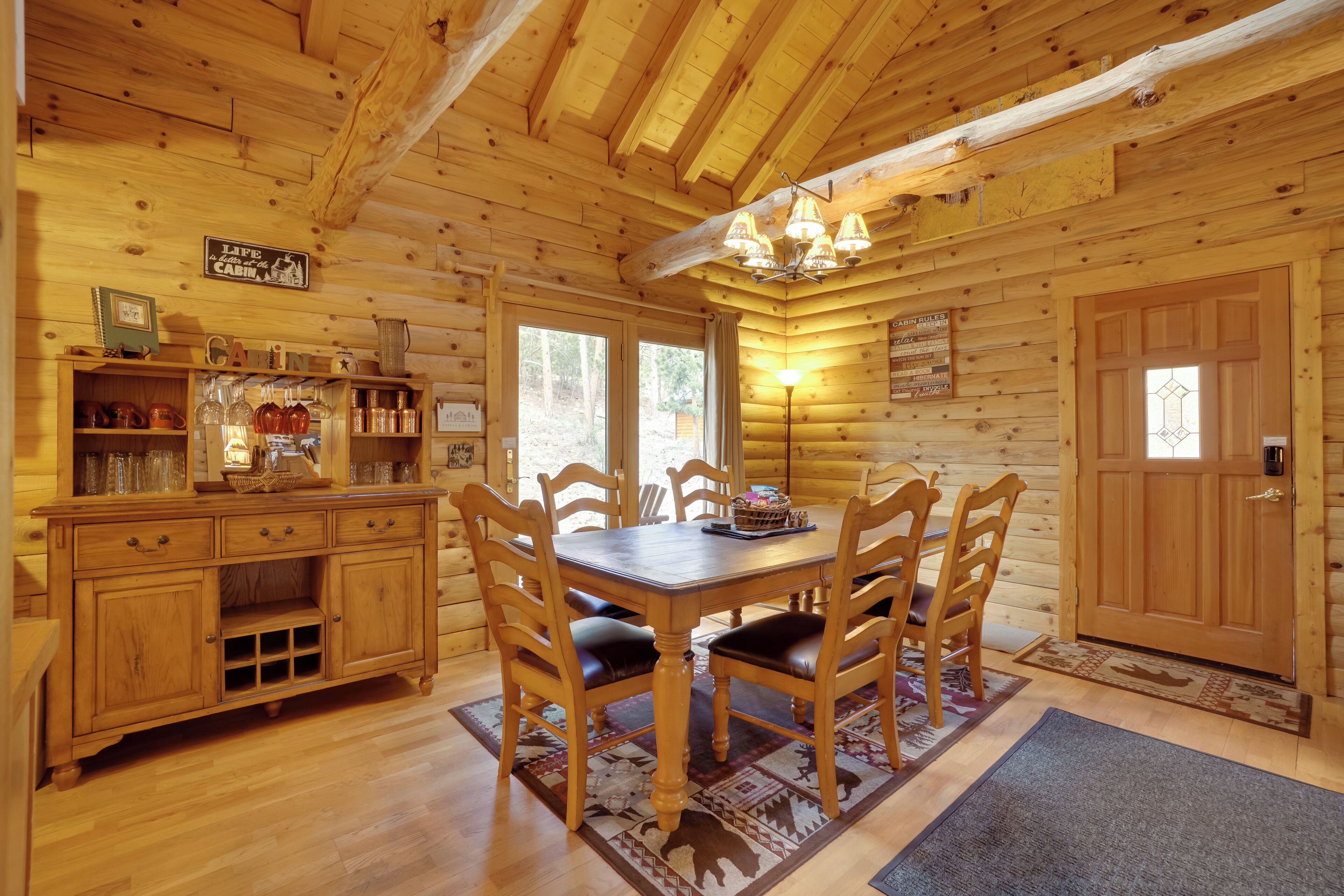 L’intérieur d’un chalet en bois comprend une table à manger, des chaises, un lustre et un coin cuisine avec des étagères et des placards.