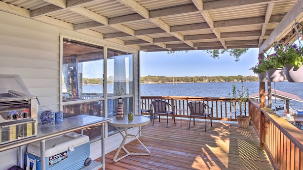 A covered outdoor deck with a grill, cooler, and seating area overlooking a body of water. - Dock & Fire Pit: Waterfront Cedar Creek Lake Home (Kemp)