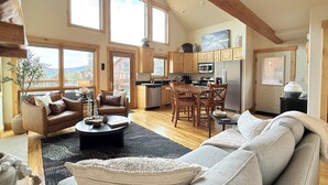A spacious living area with a large sofa, a coffee table, and a dining table with chairs. There is a kitchen area with wooden cabinets and a refrigerator.