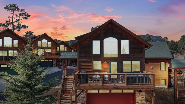 A large wooden cabin with multiple windows and a balcony, surrounded by trees and a mountain backdrop.