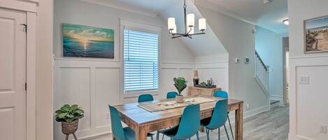 A dining area with a wooden table, teal chairs, and a chandelier. There are potted plants and a painting on the walls.