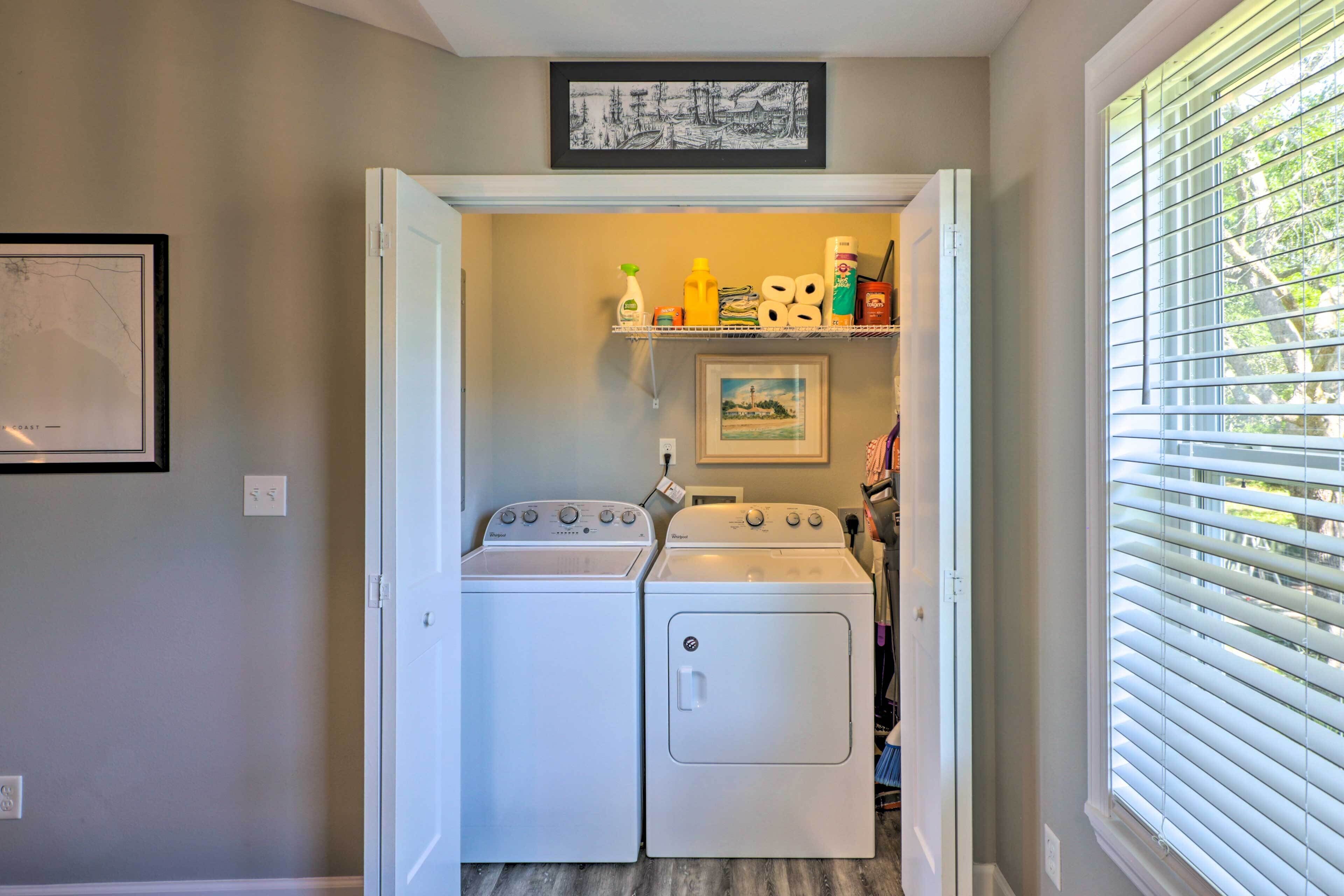 A laundry room with two white washers and dryers, a shelf with cleaning supplies, and a framed picture on the wall.